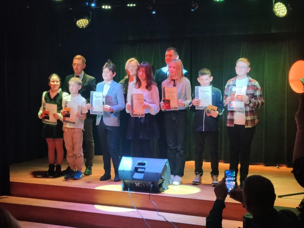 Grupa dzieci i młodzieży stojących na scenie podczas uroczystości konkursowej, trzymających dyplomy. W tle opiekunowie i organizatorzy, oświetlenie sceniczne oraz kurtyna.