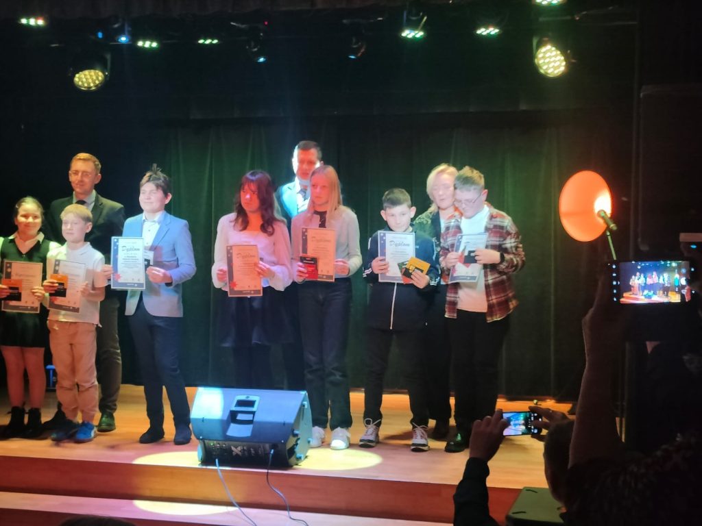 Uczniowie nagrodzeni w konkursie literackim stojący na scenie i prezentujący dyplomy. Obok nich dorośli opiekunowie, w tle zielona kurtyna i oświetlenie sceniczne.