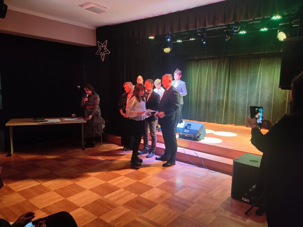 Moment wręczenia dyplomu uczennicy przez organizatora podczas uroczystości konkursowej na scenie. Obok stoją pozostali laureaci i nauczyciele.