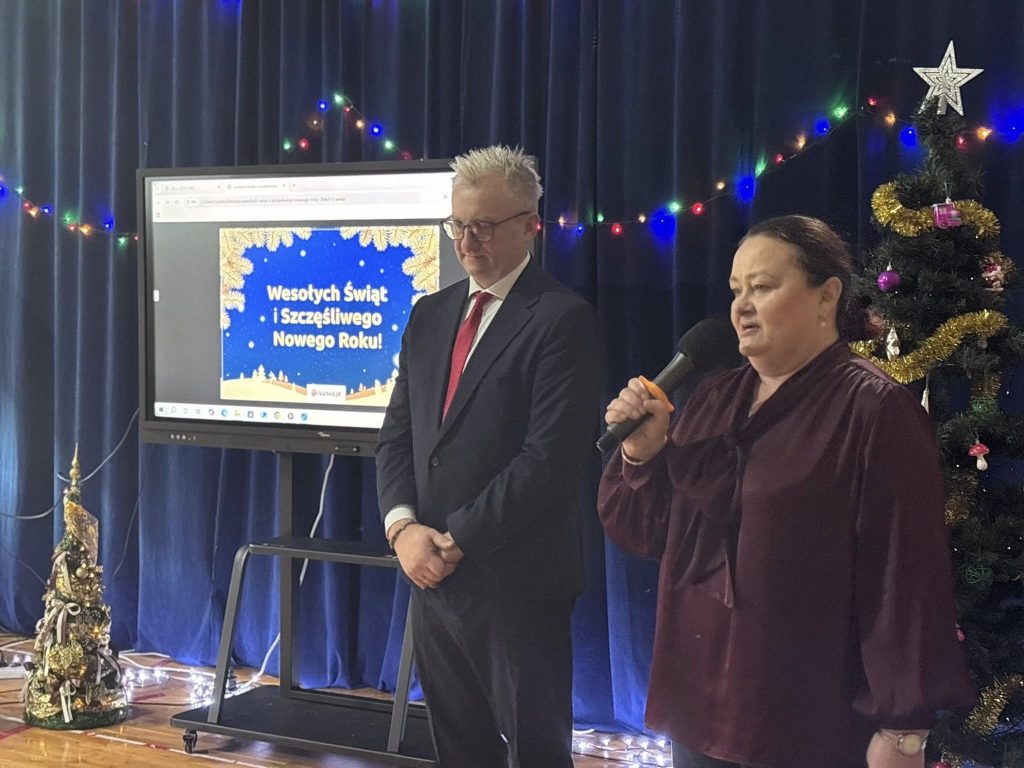 Dyrektor szkoły oraz nauczycielka stoją na scenie w świątecznej scenerii. Jedna z osób trzyma mikrofon i przemawia do zgromadzonych. W tle ekran z życzeniami świątecznymi oraz udekorowana choinka.