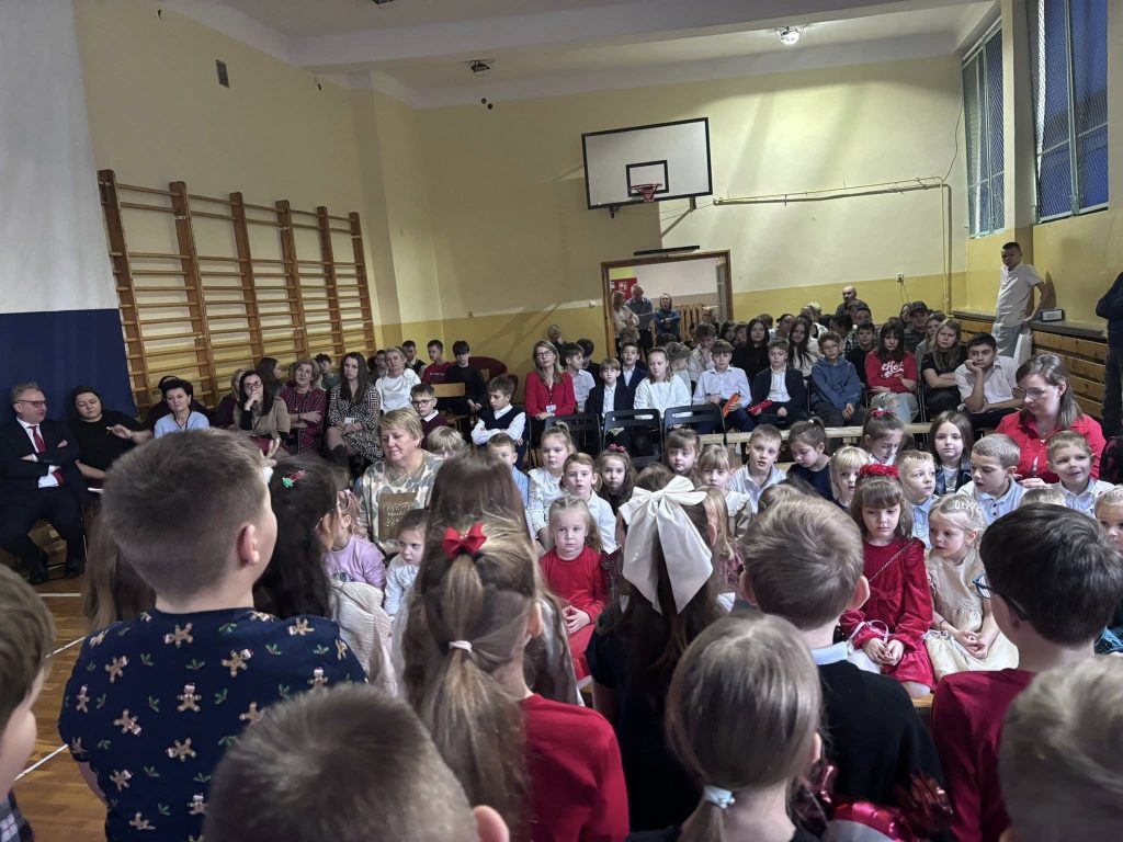 Dzieci zgromadzone na sali gimnastycznej podczas szkolnego wydarzenia świątecznego. Na pierwszym planie uczniowie, w tle nauczyciele i rodzice obserwujący występy.