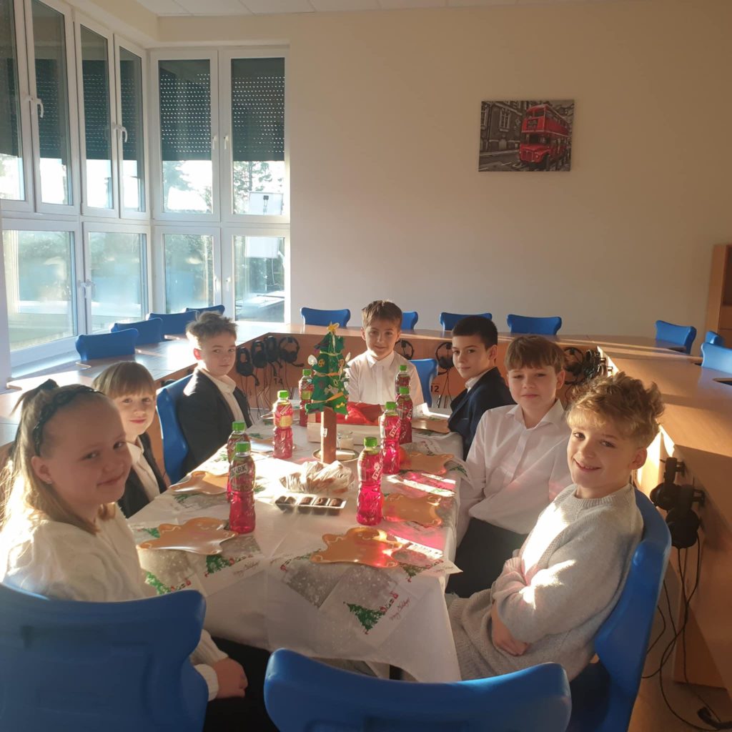 Uczniowie młodszych klas siedzą przy wspólnym stole udekorowanym świątecznie. Na stole znajdują się napoje, słodkości i dekoracje bożonarodzeniowe.