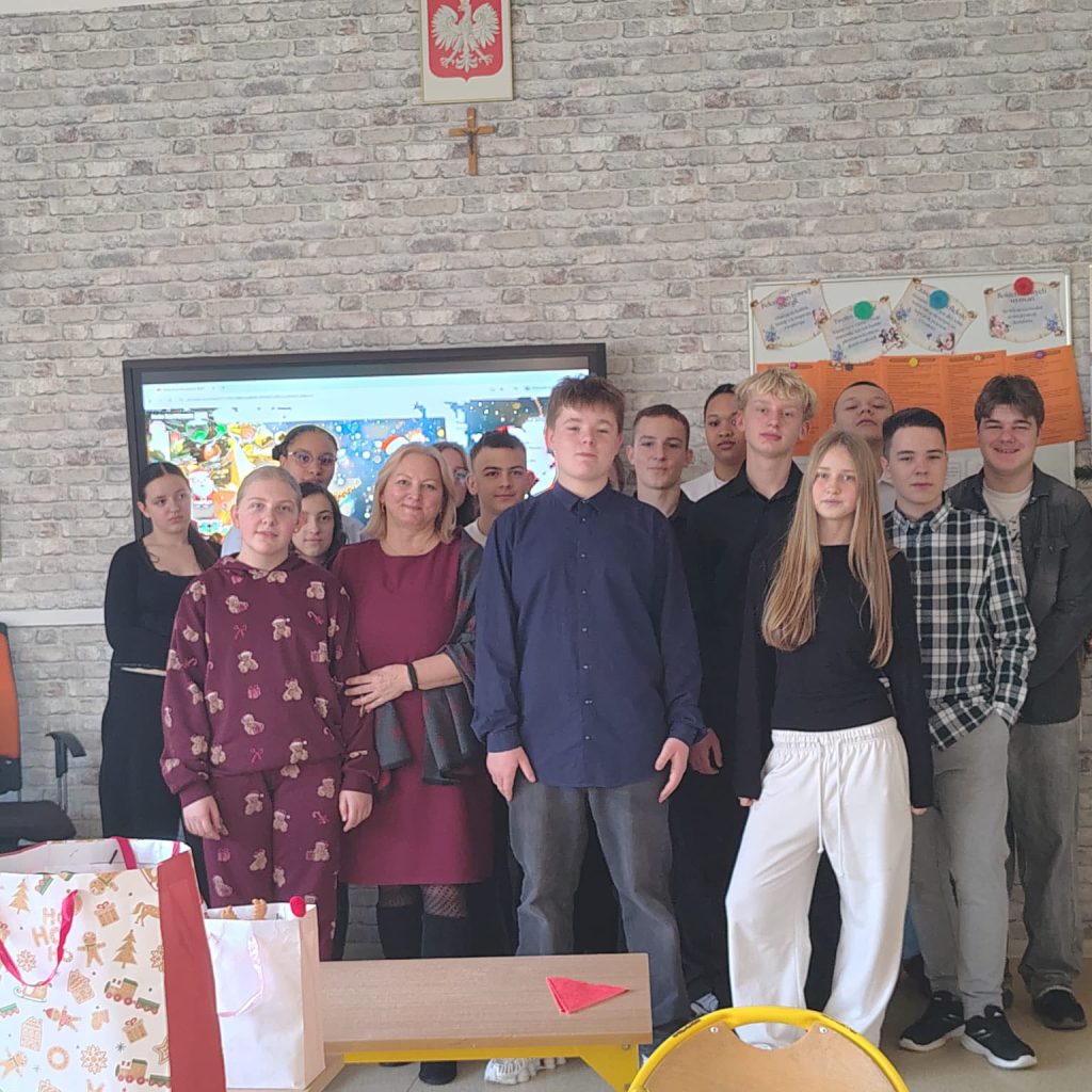 Grupa starszych uczniów wraz z nauczycielem pozuje do zdjęcia w sali lekcyjnej. W tle tablica i dekoracje świąteczne.