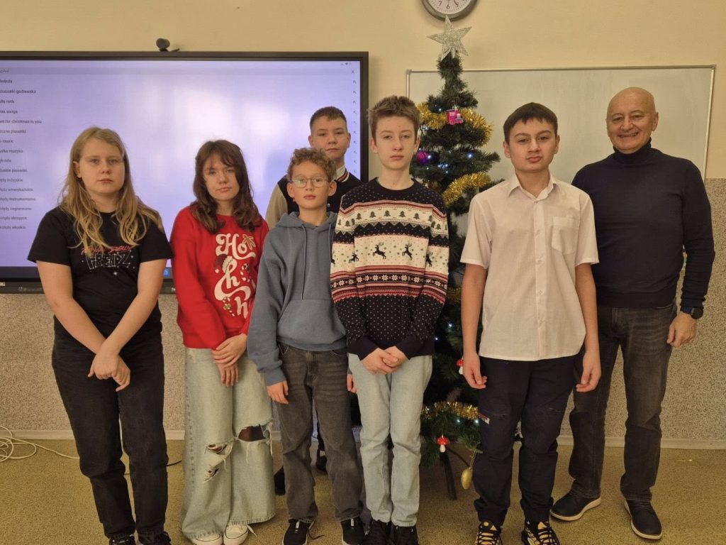 Uczniowie stojący w sali lekcyjnej obok udekorowanej choinki. Wszyscy ustawieni są do wspólnego zdjęcia w świątecznej atmosferze.