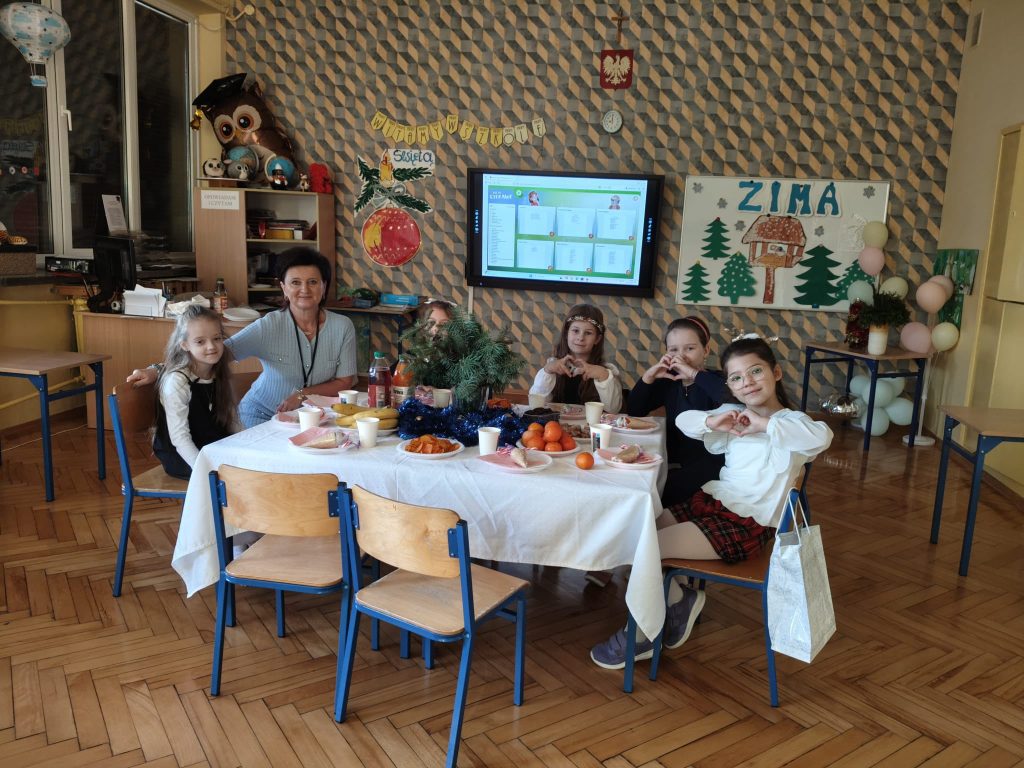 Uczniowie i nauczyciele siedzą przy wspólnym stole z przygotowanym świątecznym poczęstunkiem. Na stole widoczne są słodkości, owoce i dekoracje.