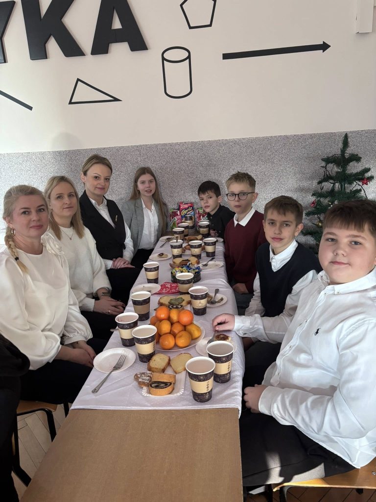 Grupa uczniów i nauczycieli zgromadzona w sali lekcyjnej podczas klasowego spotkania świątecznego. W tle dekoracje nawiązujące do zimy i Świąt Bożego Narodzenia.