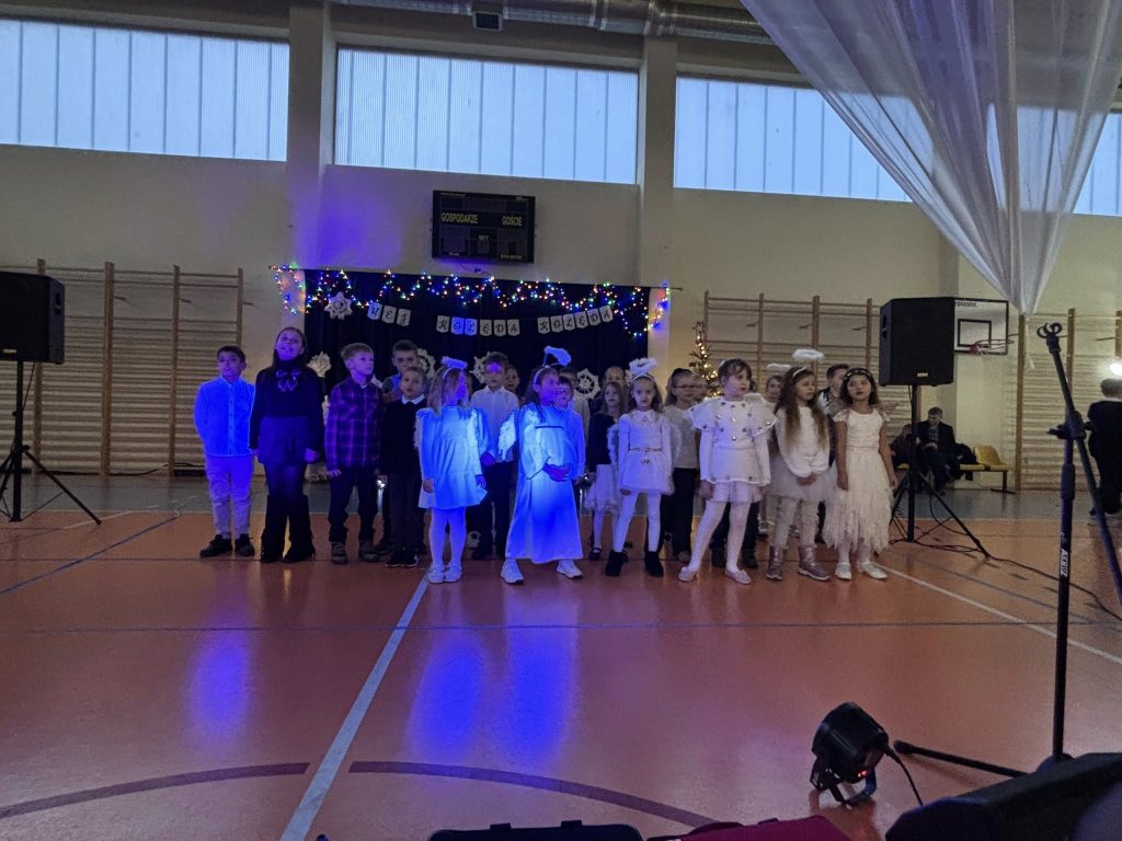 Uczestnicy Kiermaszu Świątecznego siedzą przy stołach ustawionych w sali gimnastycznej. Na stołach znajdują się dekoracje i upominki, w tle widoczni są kolejni goście oraz stoiska.