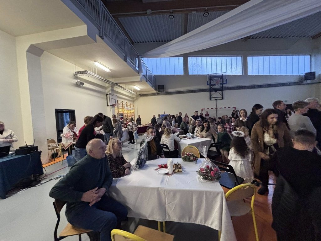 Grupa dzieci występujących na scenie w sali gimnastycznej podczas Kiermaszu Świątecznego. Uczniowie ubrani są w stroje sceniczne, a w tle widoczna jest świąteczna dekoracja.