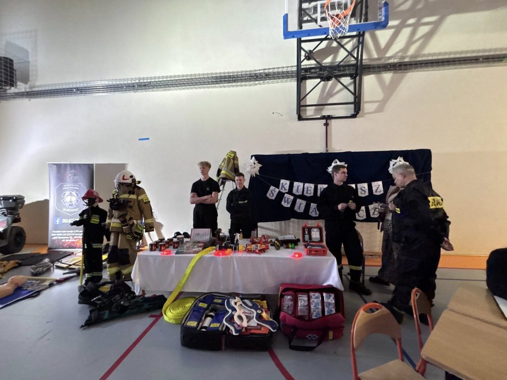 Najmłodsi uczniowie ustawieni na scenie w jasnych, świątecznych strojach podczas występu artystycznego. W tle dekoracje bożonarodzeniowe oraz oświetlenie sceniczne.