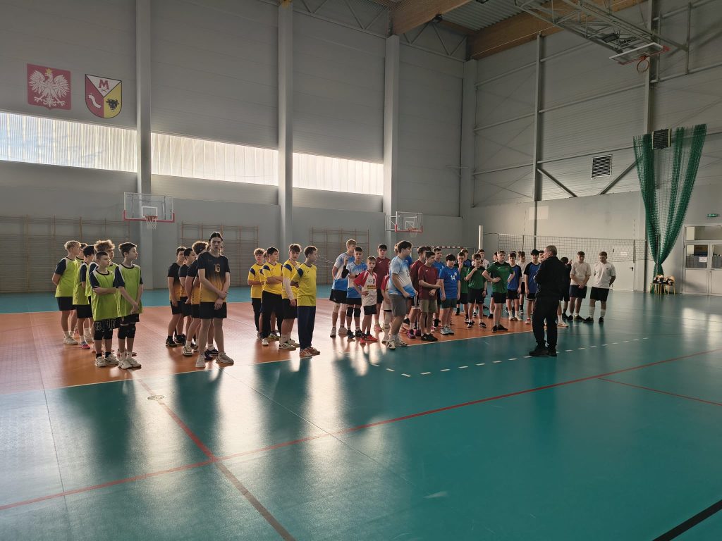 Uczniowie kilku szkół ustawieni w rzędzie na hali sportowej podczas zakończenia turnieju piłki siatkowej, nauczyciel lub organizator zwraca się do zawodników.