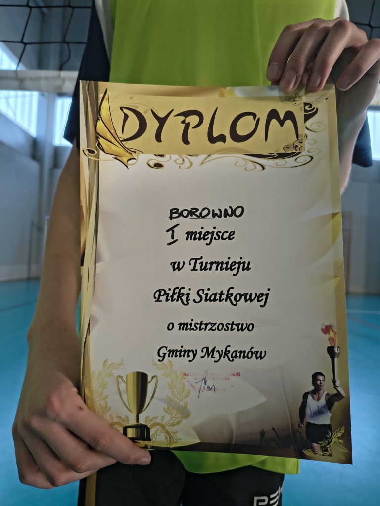 Zbliżenie na dyplom za pierwsze miejsce w turnieju piłki siatkowej o mistrzostwo Gminy Mykanów, trzymany przez ucznia w stroju sportowym na hali.