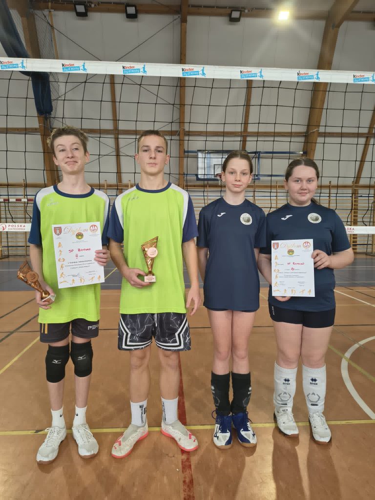 Czworo młodych zawodników stojących na hali sportowej przed siatką do piłki siatkowej. Dwoje z nich trzyma dyplomy oraz puchary zdobyte podczas turnieju.