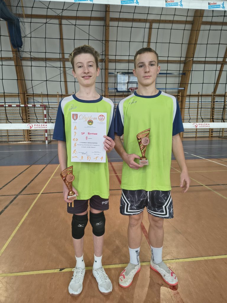 Dwóch młodych siatkarzy w zielonych strojach sportowych stojących na hali sportowej. Jeden z nich trzyma dyplom, drugi puchar za zajęcie trzeciego miejsca w turnieju.