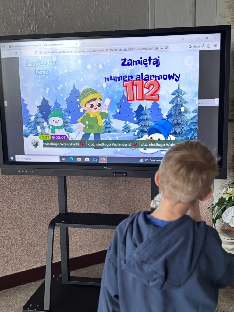 Dziecko stoi przed ekranem, na którym wyświetlona jest grafika przypominająca o numerze alarmowym 112.