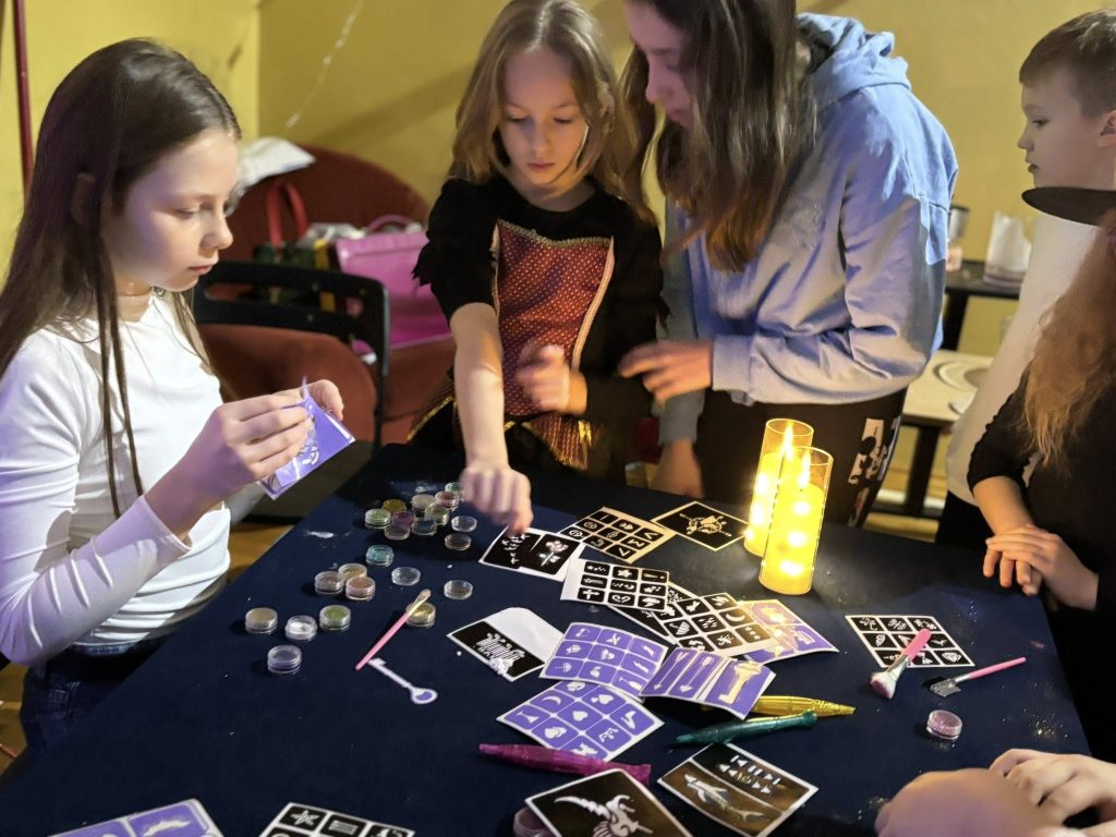 Dzieci przy stole wykonują andrzejkowe wróżby i zabawy; na blacie leżą karty, brokaty i drobne akcesoria, obok świecą lampki.