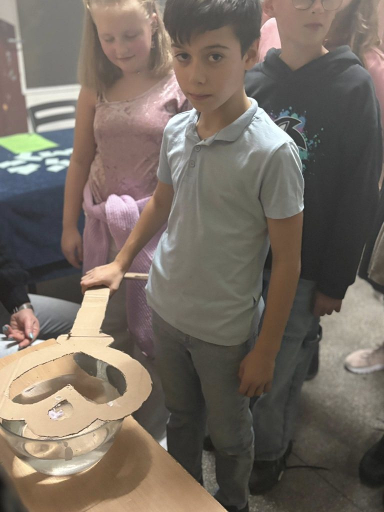 Dziecko podczas zabawy andrzejkowej stoi przy stole z rekwizytem z kartonu i miską; w tle inni uczniowie obserwują.