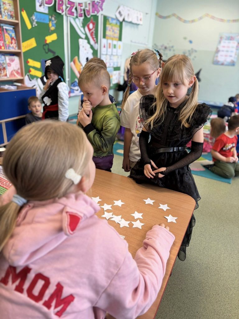 Andrzejkowa gra w klasie: dzieci stoją przy stoliku z papierowymi gwiazdkami, jedna osoba prowadzi zabawę, w tle inni uczniowie siedzą na dywanie.