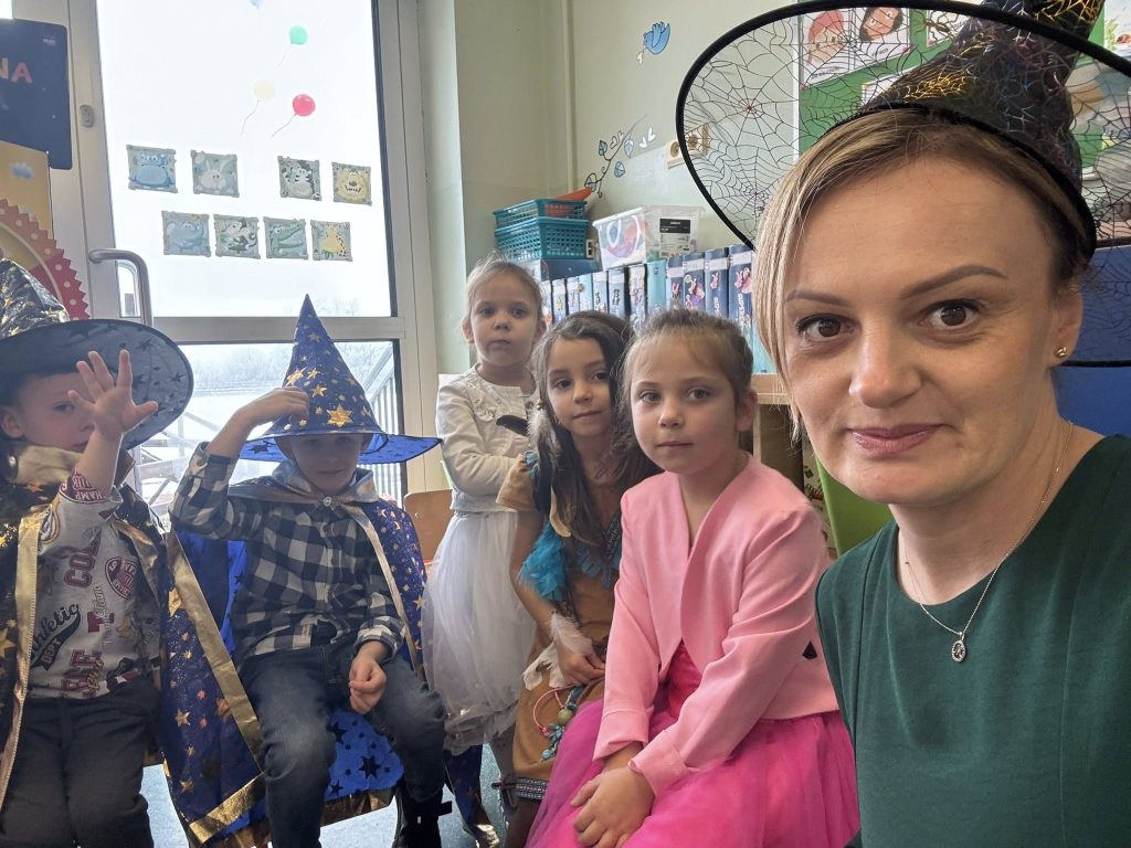 Zdjęcie w klasie podczas andrzejek: nauczycielka w kapeluszu czarownicy z dziećmi w przebraniach, część dzieci ma stroje czarodziejów i księżniczek.