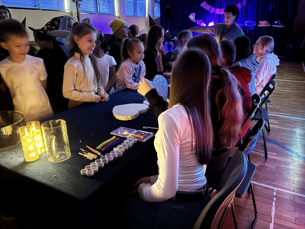 Zabawa andrzejkowa w sali gimnastycznej: dzieci ustawiają się przy stole z materiałami plastycznymi i rekwizytami, w tle widać scenę i oświetlenie.