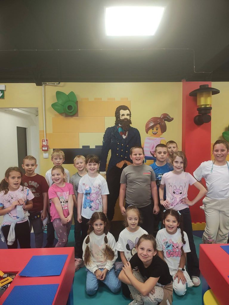 Grupa dzieci pozujących do wspólnego zdjęcia w sali edukacyjnej. W tle kolorowa ściana z figurami z klocków LEGO przedstawiającymi postacie dorosłych.
