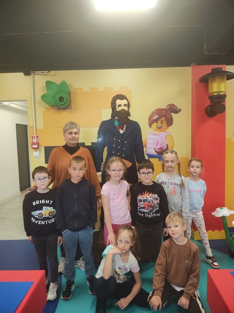 Dzieci wraz z osobą dorosłą pozują do zdjęcia w sali edukacyjnej. Za nimi dekoracyjna ściana z postaciami zbudowanymi z klocków LEGO.