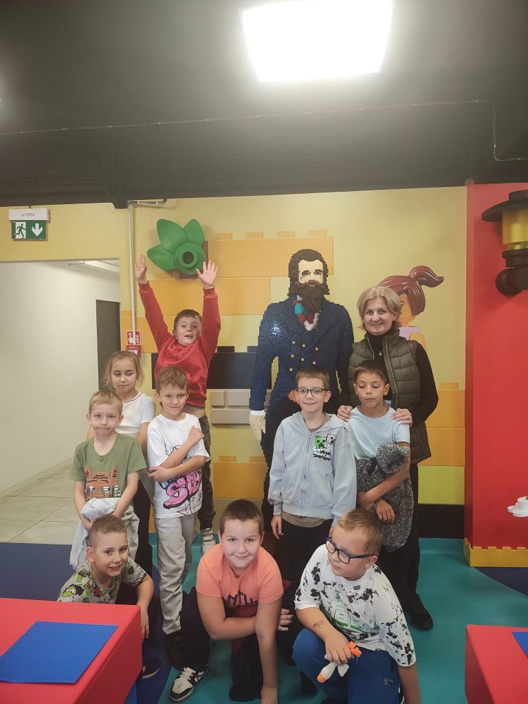 Grupa dzieci i osoba dorosła pozują do zdjęcia w sali edukacyjnej. Jedno z dzieci podnosi ręce do góry. W tle widoczna ściana z figurami z klocków LEGO.