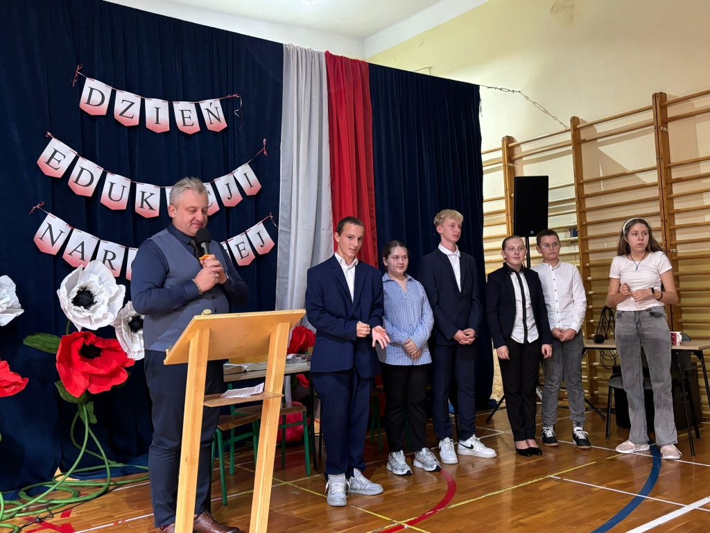 Dzień Edukacji Narodowej: mężczyzna przemawia przy mównicy, obok stoją uczniowie i prowadząca, w tle dekoracja sceniczna.
