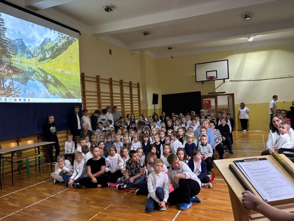 Dzień Edukacji Narodowej: uczniowie siedzą na podłodze w sali gimnastycznej i oglądają prezentację wyświetlaną na ekranie.