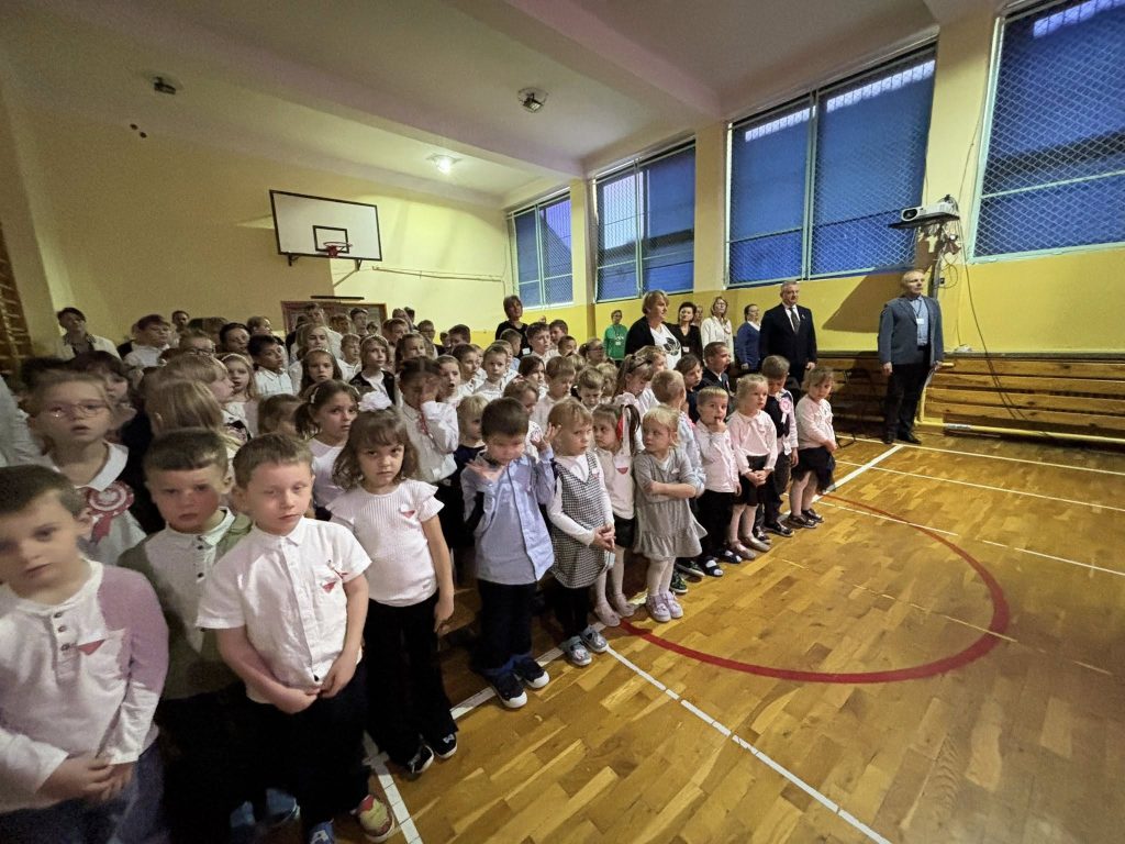Uczniowie zebrani w sali gimnastycznej podczas uroczystości szkolnej; dzieci stoją w rzędach, a z boku widać nauczycieli i gości.