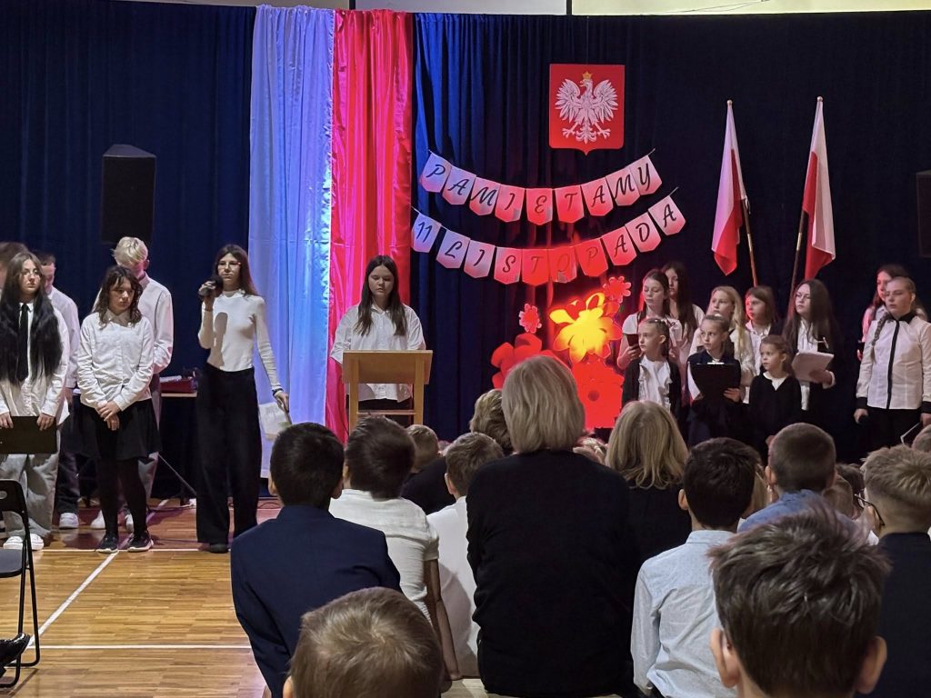 Akademia z okazji 11 listopada w sali gimnastycznej: uczniowie stoją na scenie przy mównicy, w tle dekoracja „Pamiętamy 11 listopada” i flagi Polski, na widowni zgromadzeni uczniowie.
