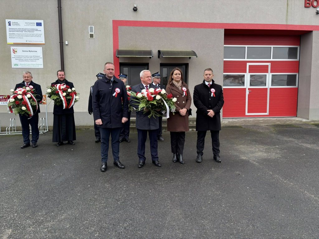 Uroczystość patriotyczna przed budynkiem straży: delegacje stoją z biało-czerwonymi wieńcami na tle czerwonej bramy garażowej.
