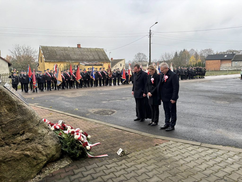 Składanie kwiatów pod pomnikiem: trzy osoby stoją przy wiązankach, w tle widać poczet sztandarowy i strażaków w szeregu.
