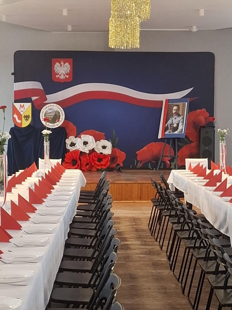 Przygotowana sala na wydarzenie okolicznościowe: dwa długie stoły nakryte białymi obrusami, biało-czerwone dekoracje i scena z tłem w barwach Polski oraz portretem.