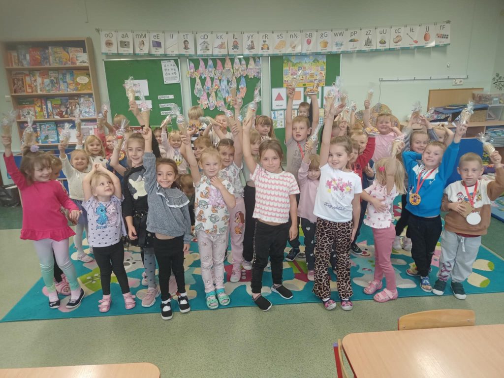 Grupa przedszkolnych dzieci stojących w sali lekcyjnej trzyma w górze drobne upominki. Na ścianach widoczne pomoce dydaktyczne i tablica.