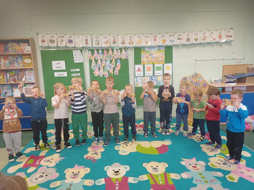 Grupa przedszkolnych dzieci ustawionych w rzędzie prezentuje otrzymane nagrody. Dzieci stoją na kolorowym dywanie w sali edukacyjnej.