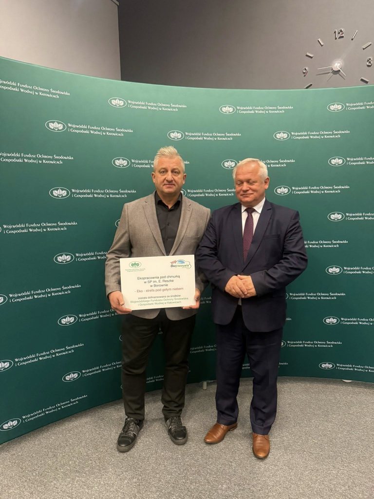 Dwóch mężczyzn stojących przed zieloną ścianką Wojewódzkiego Funduszu Ochrony Środowiska i Gospodarki Wodnej w Katowicach. Jeden z nich trzyma certyfikat potwierdzający realizację projektu ekologicznego.
