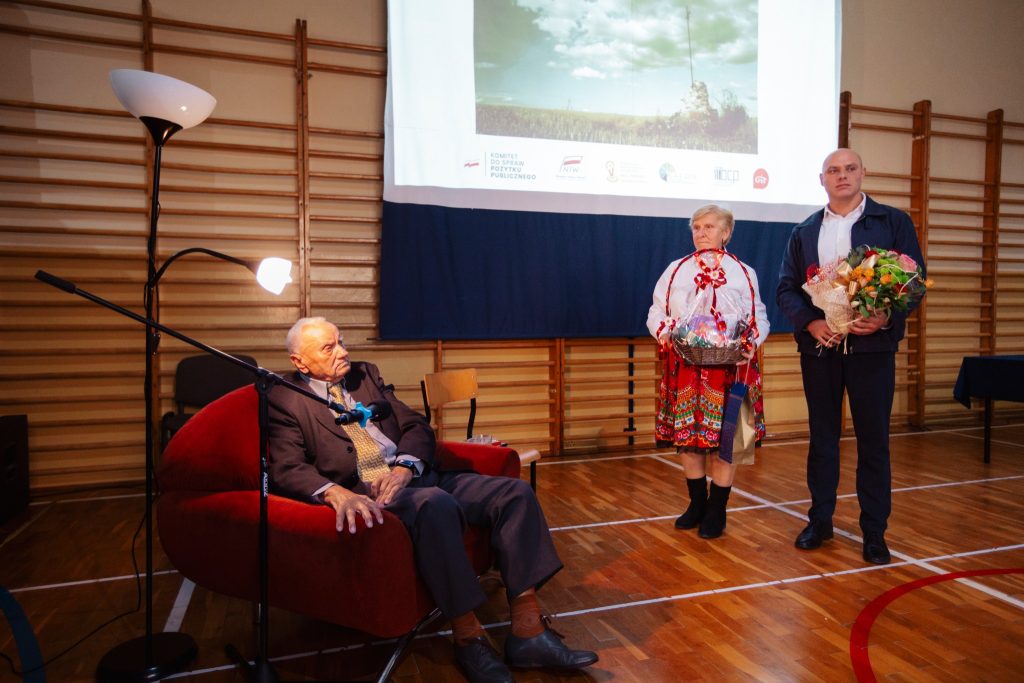 Uhonorowanie seniora podczas wydarzenia „Moja miejscowość wczoraj i dziś”: starszy mężczyzna siedzi na fotelu, obok stoją dwie osoby z kwiatami i upominkiem.