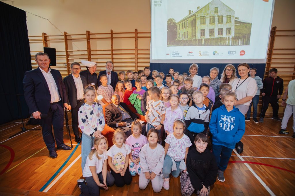 Zdjęcie grupowe uczniów i dorosłych w sali gimnastycznej podczas wydarzenia „Moja miejscowość wczoraj i dziś”, z ekranem prezentacji w tle.
