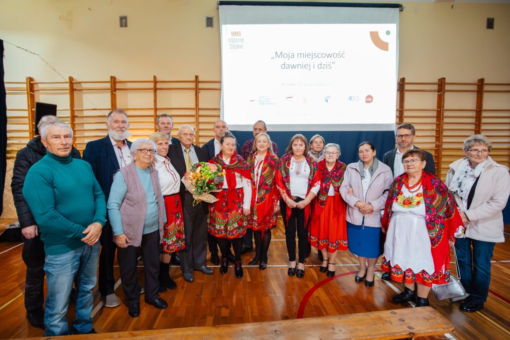 Uczestnicy wydarzenia „Moja miejscowość wczoraj i dziś” pozują do zdjęcia w sali gimnastycznej; część osób ma stroje ludowe, w tle ekran prezentacji.