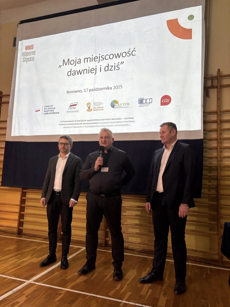 Trzej prowadzący stoją przed ekranem z tytułem „Moja miejscowość dawniej i dziś” podczas spotkania w sali gimnastycznej.
