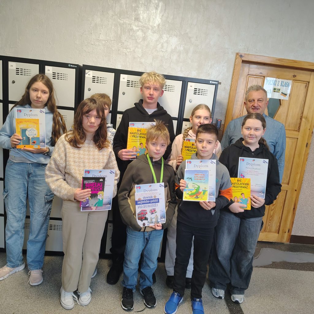 Grupa uczniów stoi w szkolnym korytarzu przy szafkach. Uczniowie trzymają dyplomy i nagrody książkowe; obok stoi dyrektor.
