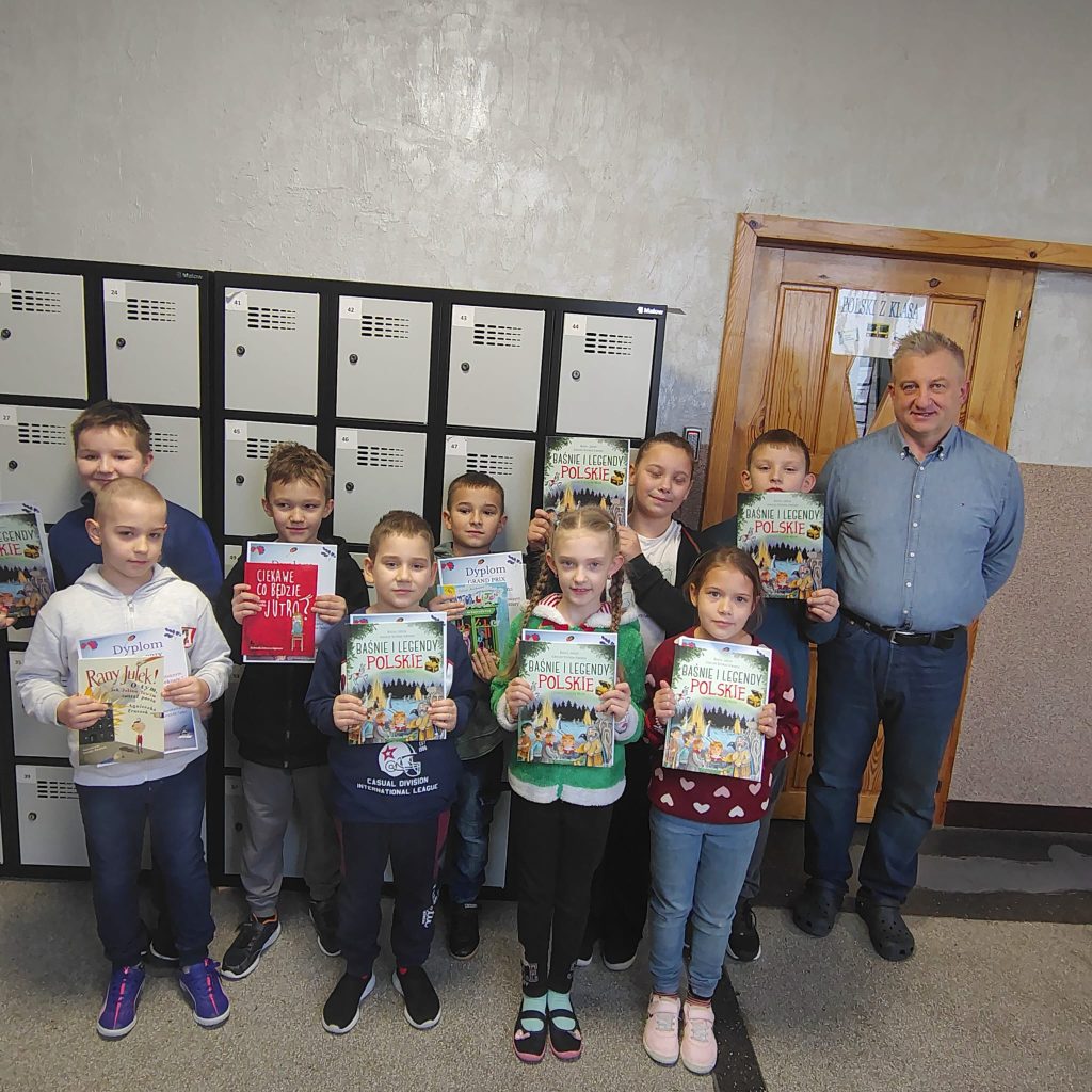 Uczniowie z dyplomami i książkami pozują w szkolnym korytarzu na tle szafek. Po prawej stronie stoi dyrektor.