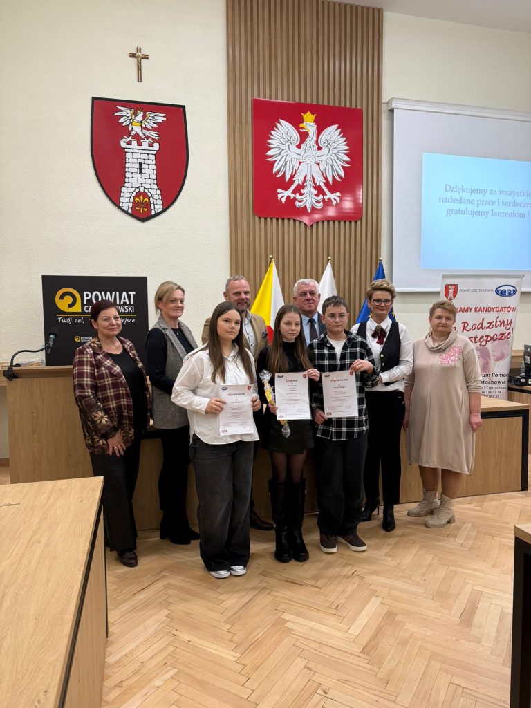 Zdjęcie grupowe z uroczystości w sali urzędu: troje uczniów stoi z dyplomami (i upominkiem), za nimi opiekunowie i organizatorzy. W tle godło Polski, flagi oraz baner powiatu.