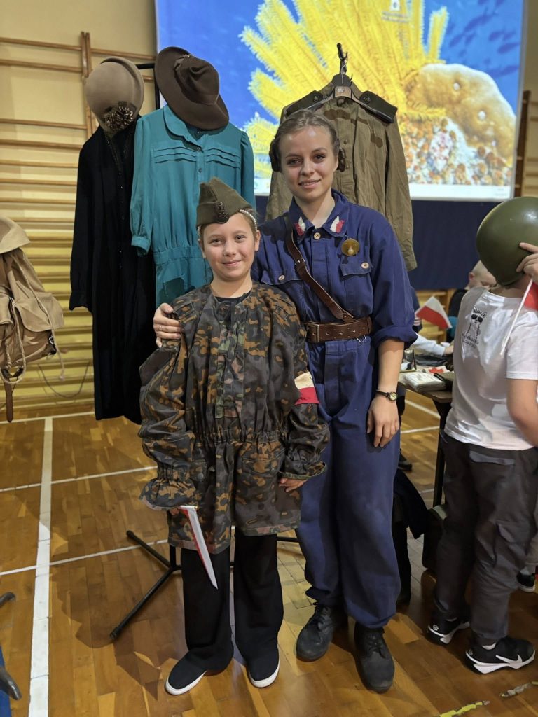 Prowadząca lekcję historyczną stoi obok dziecka ubranego w historyczny mundur wojskowy. Oboje pozują do zdjęcia na tle ekspozycji z umundurowaniem i elementami wyposażenia z okresu II wojny światowej.