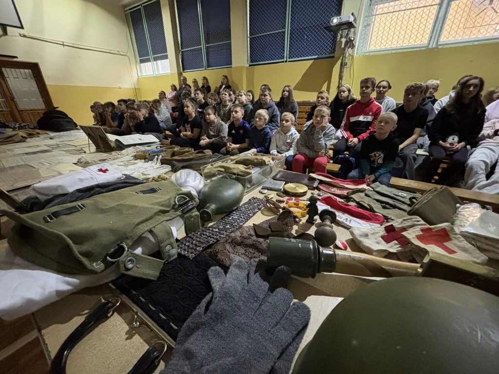 Grupa dzieci siedzących na ławkach w sali gimnastycznej słucha prelekcji historycznej. Na pierwszym planie stoły z eksponatami z okresu II wojny światowej, w tym hełmy, maska przeciwgazowa, opaski Czerwonego Krzyża i elementy umundurowania.