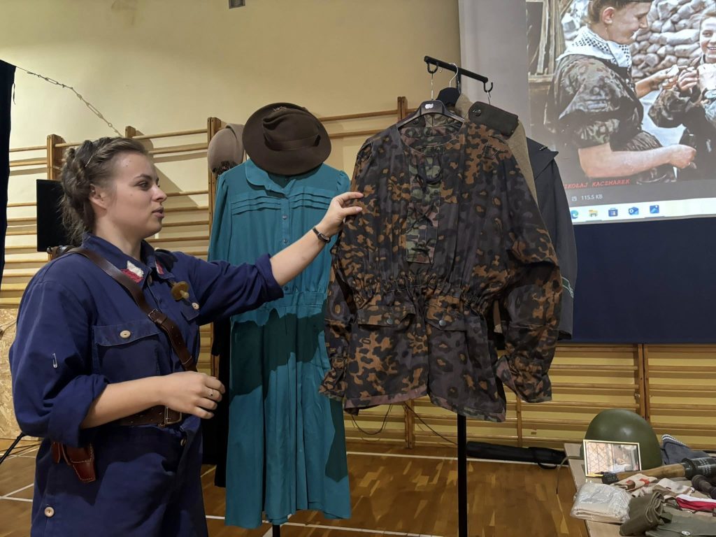 Prowadząca prelekcję pokazuje dziecięcy mundur wojskowy w kamuflażu z okresu II wojny światowej. Obok na stojakach wiszą historyczne ubrania cywilne i wojskowe.