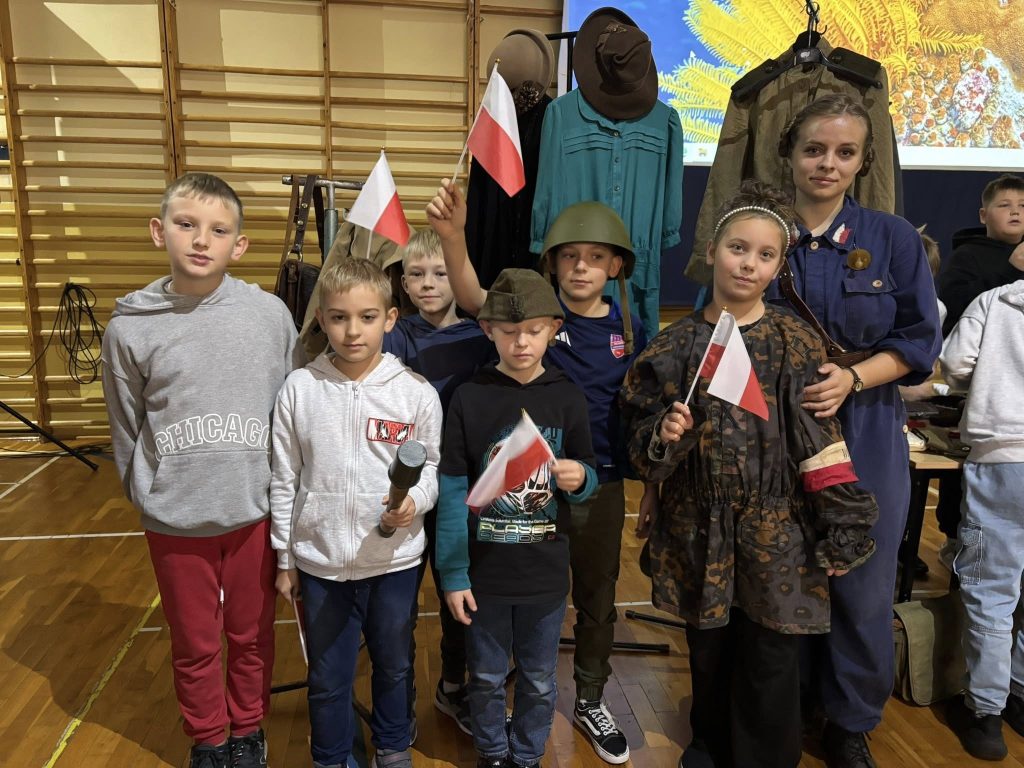 Grupa dzieci pozujących do zdjęcia po lekcji historycznej. Część dzieci trzyma biało-czerwone chorągiewki, jedno z nich ma na sobie hełm wojskowy z okresu II wojny światowej. W tle widoczne historyczne stroje na stojakach.