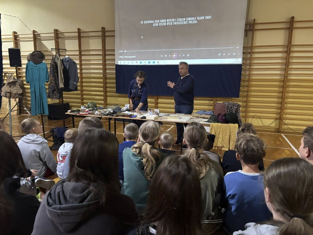 Widok od strony publiczności na lekcję historyczną w sali gimnastycznej. Dwoje prowadzących stoi przy stołach z eksponatami, a na ekranie wyświetlany jest slajd z informacją o wydarzeniach z 1939 roku.