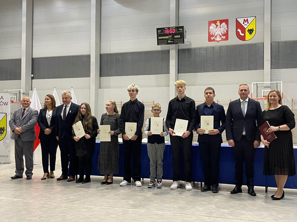 Uroczystość w hali sportowej: grupa uczniów z dyplomami stoi w szeregu razem z przedstawicielami gminy. Zdjęcie z wręczenia Nagrody Wójta Gminy Mykanów.