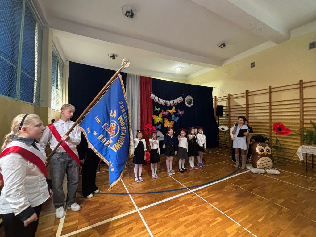 Grupowe zdjęcie uczniów w biretach z nauczycielami i rodzicami na tle dekoracji „Ślubujemy” w sali gimnastycznej.